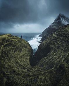 a man standing on top of a lush green hillside next to the ocean under a cloudy sky