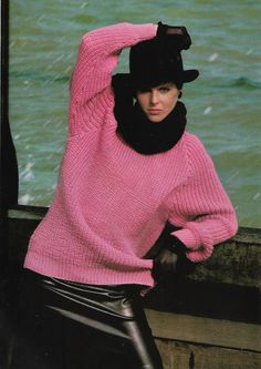 a woman in a pink sweater and black hat leaning against a wall near the water