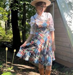 a woman wearing a dress and hat standing in front of a shed with flowers on the ground