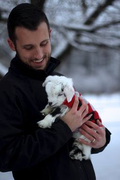 a man holding a white and black dog in his arms while standing next to snow covered trees