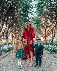 a woman in a red dress and two children are walking down a brick path lined with trees