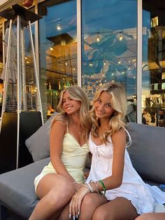 two beautiful young women sitting next to each other on top of a gray couch in front of a window