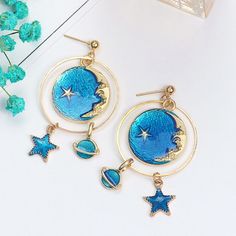 "Beautifully designed earrings featuring a moon and celestial theme with lapis-blue colors. The earrings are drop/dangle style hook. They will drop down 3\". One half of the set features a Saturn-like planet charm within an open metal circle, and a star at the bottom. The planet charm measures approximately 0.5\" in diameter, and the width of the star is approximately 0.3\". The other half of the set features a star at the top, then a round charm near the base. The round charm measures approxima Blue Dangle Hoop Earrings, Blue Round Earrings With Moon Charm, Blue Moon-shaped Metal Jewelry, Blue Metal Dangle Hoop Earrings, Blue Moon Shaped Earrings With Moon Charm, Blue Moon-shaped Earrings With Moon Charm, Blue Celestial Pierced Earrings, Celestial Blue Pierced Earrings, Blue Dangle Earrings With Moon Charm