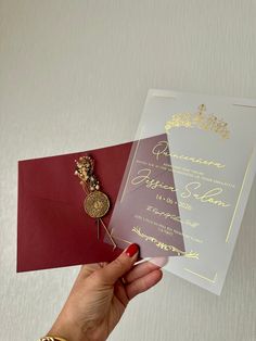 a person holding up a red and white wedding card with a gold brooch on it