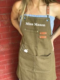 a woman standing next to a red brick wall wearing an apron with the name miss mason on it