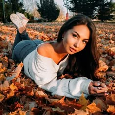 a beautiful woman laying on the ground covered in autumn leaves and looking at the camera