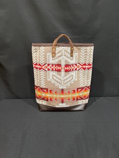 This is a tall book bag with short leather handles. The pattern is in a tan and white and is handmade using Pendleton® wool and good quality technical fabric. It has a very roomy interior that is lined with a natural-colored heavy cotton canvas and has a magnetic closure and a hanging interior zippered pocket. The bag has short, black leather handles and the top is bound with brown faux suede. The fabric pattern is in tan and white (with some red and orange) and tech fabric on the bottom is brow Wool Tote Bag For Travel, Rectangular Wool Shoulder Bag For Travel, Wool Rectangular Shoulder Bag For Travel, Wool Tote Bag For Daily Use, Rectangular Wool Travel Bag, Daily Use Wool Tote Bag, Wool Tote Bag For Everyday Use, Daily Use Wool Bags With Leather Handles, Daily Use Wool Bag With Leather Handles