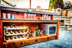 an outdoor bar with wine bottles and glasses on the shelves, next to a mini fridge