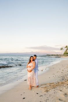 a man and woman standing on top of a sandy beach next to the ocean at sunset