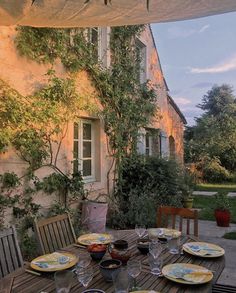 an outdoor dining table with plates and glasses on it, in front of a house