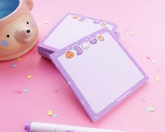 a notepad and pen sitting next to a cup on a pink surface with confetti