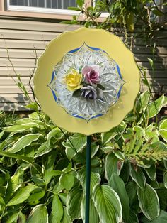 a yellow plate with flowers in it sitting on top of a green pole next to some bushes