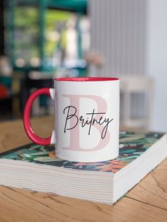 a red and white coffee mug sitting on top of a wooden table next to a book