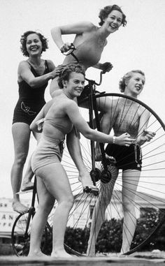 three women in bathing suits are standing next to an old bicycle and one woman is holding the wheel