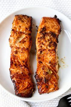 two pieces of fish on a white plate next to a fork and knife with another piece of meat in the background