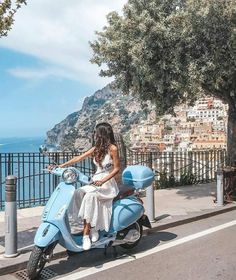 a woman is sitting on a blue scooter by the water and trees in front of her