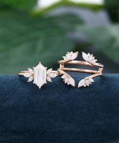 two gold rings with white stones and leaves on the top, sitting on a blue velvet surface