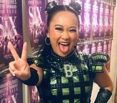 a woman making the peace sign in front of a wall with posters behind her on it