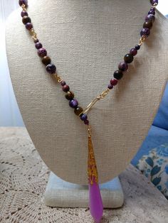 A lariat necklace made of purple jasper and tiger's eye beads on a gold-plated chain, with a purple and white resin pendant in a gold-plated iron setting. The necklace is 25 3/4 inches long from end to end and the pendant glows blue in the dark if exposed to UV light. To where it, the pendant goes through the gold-plated loop on the opposite end, and it can be tightened to the wearer's liking. Adjustable Gold Lariat Necklace With Natural Stones, Unique Purple Dangle Necklaces, Gold Lariat Necklace With Natural Stones, Gold Teardrop Agate Necklace, Gold Bohemian Lariat Necklace With Natural Stones, Bohemian Gold Lariat Necklace With Gemstone Beads, Gold Bohemian Lariat Necklace With Gemstone Beads, Handmade Purple Lariat Necklace, Gift Natural Stones Dangle Lariat Necklace