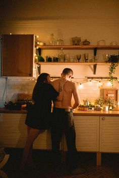 a man and woman standing in front of a kitchen counter with candles on the shelves