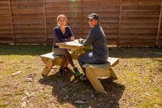 a man and woman sitting at a picnic table in front of a fenced yard