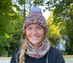 a woman wearing a knitted hat and scarf with a fur pom in front of trees