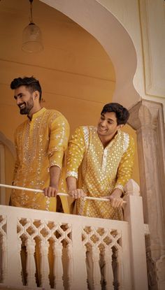 two men dressed in yellow standing on a balcony with white railings and an archway behind them