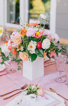 an arrangement of flowers in a vase on a pink table cloth with place settings and utensils