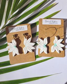 two white flower shaped earrings sitting on top of a piece of cardboard next to a palm leaf