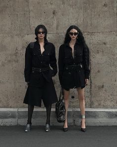 two women standing next to each other in front of a concrete wall with sunglasses on