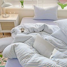 an unmade bed with blue and white striped comforter on it, next to a window