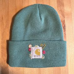 a green beanie with an embroidered patch on the front and side, sitting on top of a wooden floor