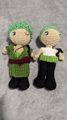 two crocheted dolls standing next to each other on a gray surface, one with green hair and the other wearing black pants