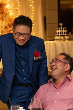 two men sitting at a table with wine glasses in front of them and one is smiling