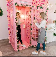 two women standing in front of a pink phone booth decorated with flowers and greenery