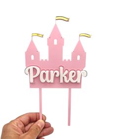 a hand holding up a pink cake topper with the word parker on it and a castle