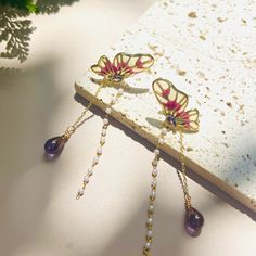 These exquisite butterfly earrings 🦋 feature delicate, golden butterflies with vibrant red and yellow wings, highlighted by a central gemstone 💎. Dangling from each butterfly is a chain of small, white beads, adding a touch of elegance 🌟. At the end of the chain, a stunning purple teardrop gemstone completes the look. These earrings are perfect for adding a whimsical and elegant flair to any outfit, making them a unique and charming addition to any jewelry collection 🌸. Handcrafted with meti Fairycore Gold Drop Earrings, Handmade Gold Fairy Earrings, Gold Handmade Fairy Earrings, Gold Fairy Earrings For Gift, Gold Fairy Style Handmade Earrings, Gold Fairycore Drop Earrings, Red Butterfly Earrings For Gift, Fairycore Butterfly Earrings For Gift, Gold Fantasy Earrings As Gift