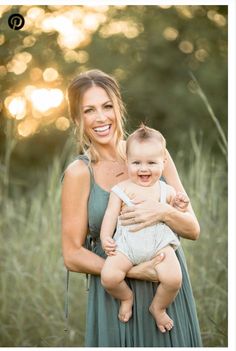 a woman holding a baby in her arms and smiling at the camera while standing in tall grass