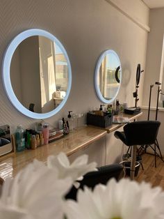 two round mirrors on the wall above a counter with flowers in front of it and a hair dryer nearby