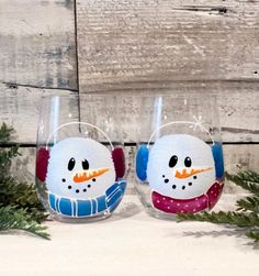 two wine glasses with snowmen painted on them sitting next to evergreen branches and wood planks