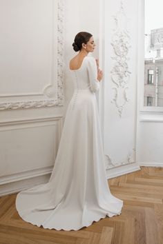 a woman in a white wedding dress is standing near a wall and looking at something