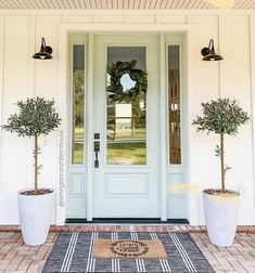 two potted trees are on the front porch with a welcome mat and doormat