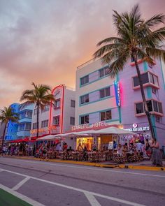 people are sitting at tables in front of a building with neon lights and palm trees