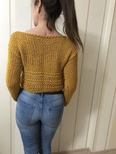 a woman standing in front of a wall wearing jeans and a yellow knitted sweater