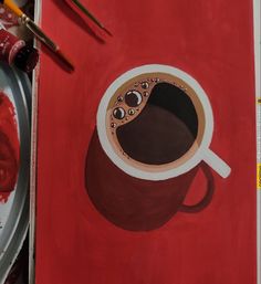 a painting of a cup of coffee on a red table with paintbrushes next to it