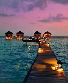 a dock leading into the water with lit up huts in the background at sunset or dawn