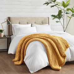 a bed with white linens and yellow blankets on it, next to a potted plant