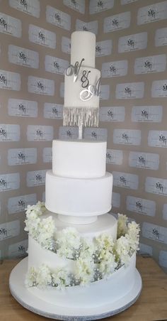 a three tiered white wedding cake with flowers on the bottom and monogrammed wallpaper behind it