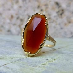 This large 10 karat gold ring features a single shiny, orange-red carnelian center stone with a bold geometric bezel setting. A beautiful piece from the late 1970's-80's. This large gem is held in place by uniquely set bezel mounting. It's a wonderfully crafted ring ring full of classic details. The carnelian cabochon measures 12.5mm by 21.6mm.  The inside of the band is not stamped but tests as 10 karat. The condition of this lovely ring is shows very light wear to the gold. The gold has been n Carnelian Ring, Lovely Ring, Ring Ring, Bezel Setting, Solitaire Ring, Orange Red, Gold Ring, Gold Rings, Jewelry Rings