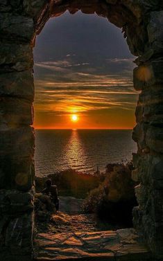 the sun is setting over the ocean through an arch in stone wall with water below